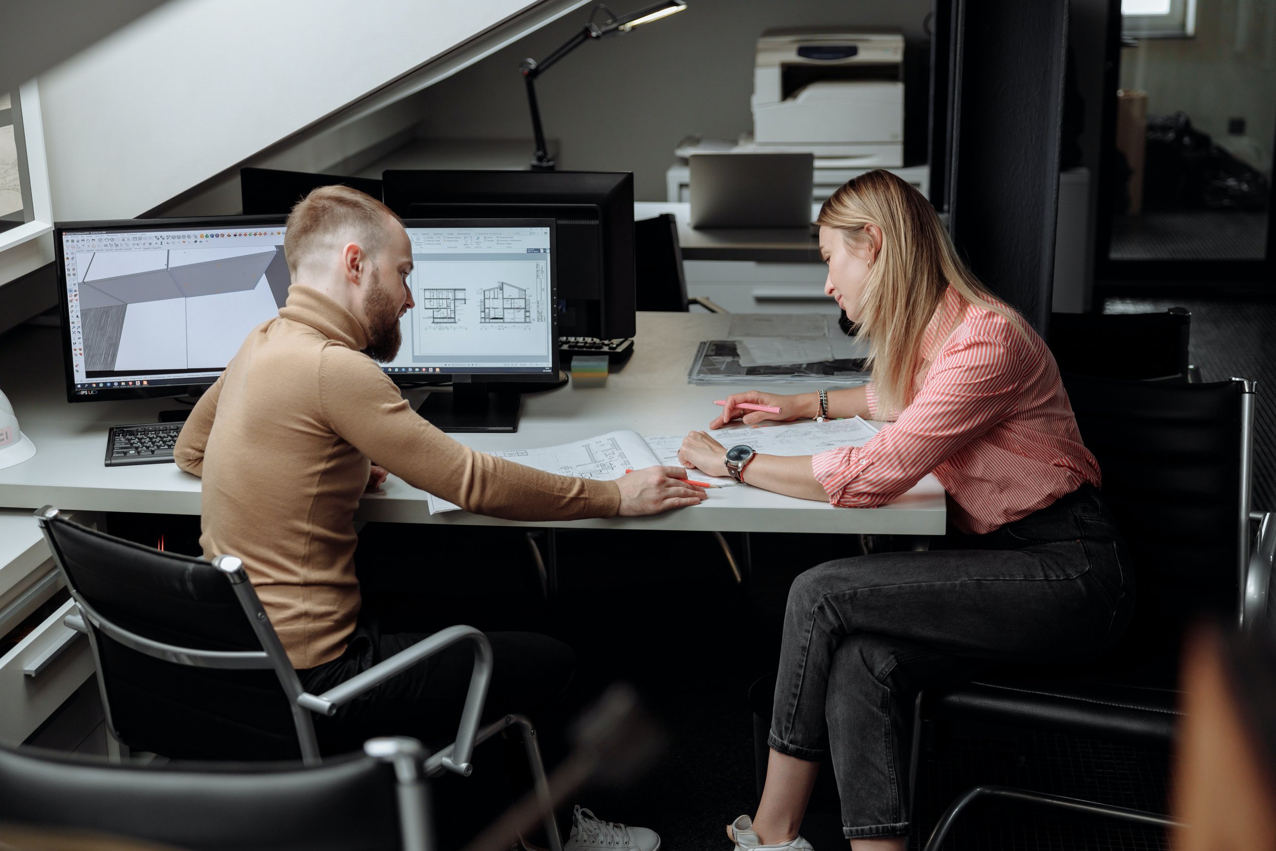 A man and a woman sitting at a desk looking at a series of blueprints printed on a table with a computer that has construction plans on the screen.