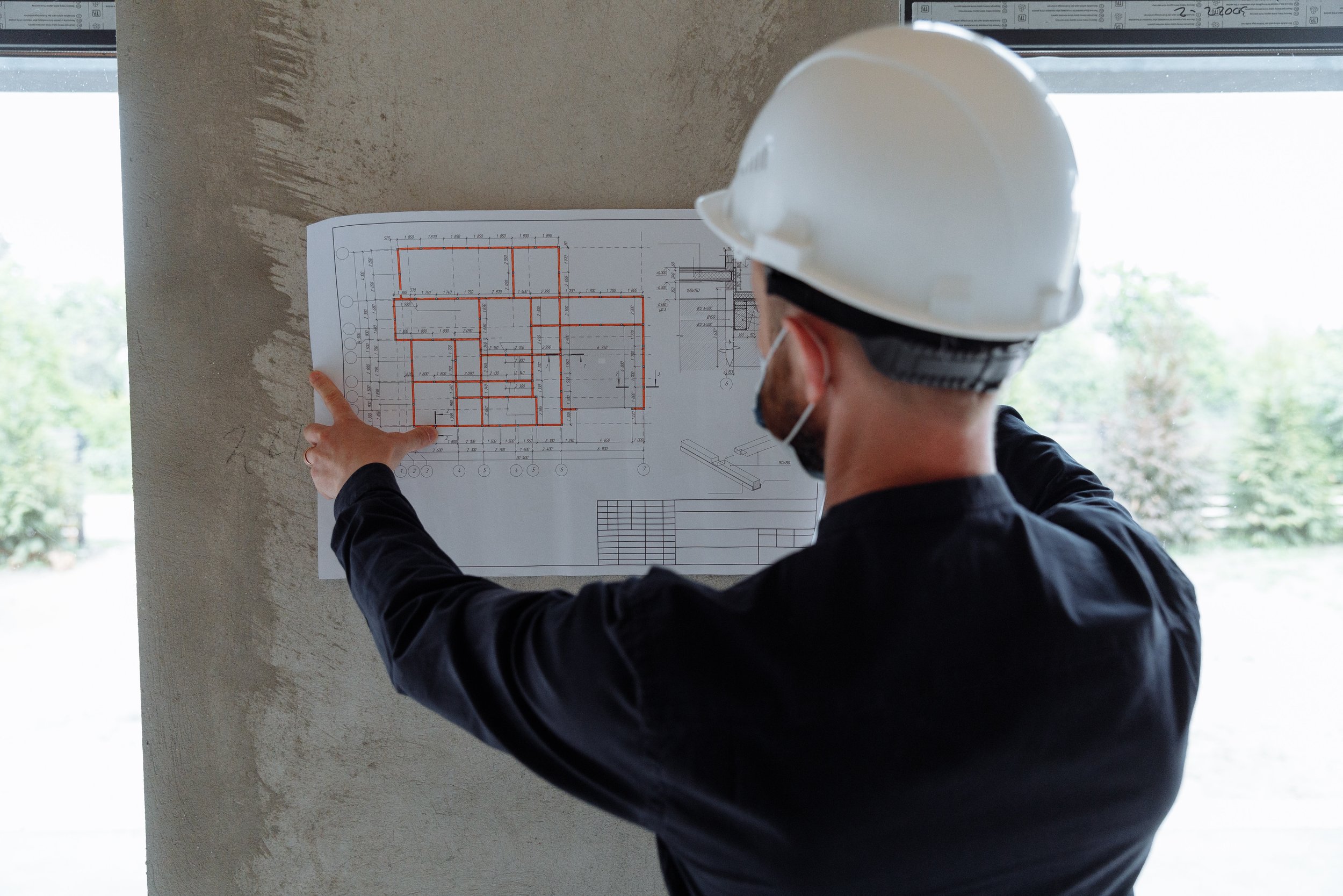 A person wearing a hard hat holding a blueprint of a house remodeling project against a wall.